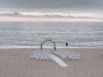 A wedding ceremony setup on a beach features an archway adorned with greenery and white flowers, surrounded by rows of white chairs arranged symmetrically facing the arch. The ocean waves gently roll in the background under a cloudy sky with a hint of sunlight breaking through the clouds along the horizon.