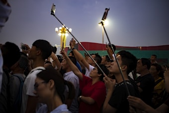 A group of people is gathered in the evening, holding up smartphones and selfie sticks to capture a view, possibly a performance or an event. The crowd is diverse, with individuals looking upwards, illuminated by artificial lights against a dusky sky.