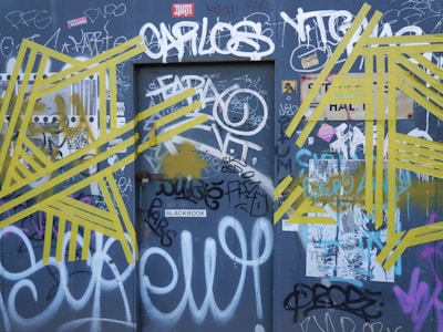 A wall covered in various graffiti and street art, featuring white, yellow, and multi-colored spray-painted tags and designs. Prominent yellow geometric patterns intersect with diverse black and white text, contributing to a chaotic and artistic urban aesthetic.