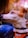 A small dog with white and brown fur is looking upward. The dog wears an orange harness with peace, heart, and paw symbols on a tag. The background is blurred with warm tones, suggesting a natural or outdoor setting. A person is partially visible, appearing to hold or pet the dog.