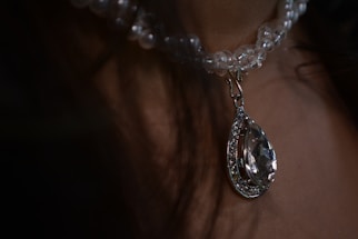 A close-up view of a clear beaded necklace featuring a large pear-shaped crystal pendant with intricate detailing, hanging against a neutral background.