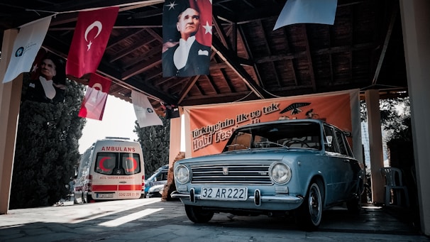 A vintage blue car is parked under a wooden-roofed structure adorned with Turkish flags and banners. An ambulance is seen parked nearby with its back visible. Portraits of a historical figure are displayed on the banners. The setting seems to be an outdoor cultural or hobby festival.