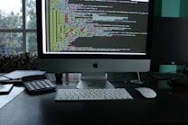 A workspace featuring a sleek desktop computer with a large screen displaying lines of colorful code. The setup also includes a keyboard, a mouse, a calculator, and a smartphone placed on the desk. The backdrop consists of a large window with a view of trees and an urban environment, suggesting a modern office setting.