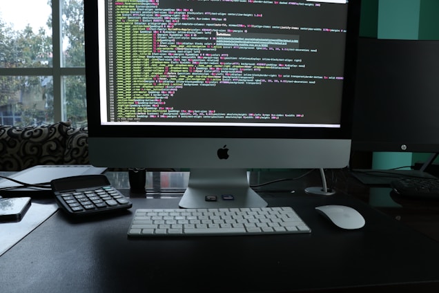 A workspace featuring a sleek desktop computer with a large screen displaying lines of colorful code. The setup also includes a keyboard, a mouse, a calculator, and a smartphone placed on the desk. The backdrop consists of a large window with a view of trees and an urban environment, suggesting a modern office setting.