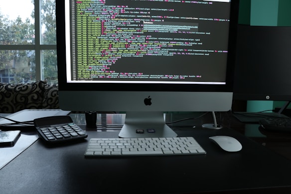 A workspace featuring a sleek desktop computer with a large screen displaying lines of colorful code. The setup also includes a keyboard, a mouse, a calculator, and a smartphone placed on the desk. The backdrop consists of a large window with a view of trees and an urban environment, suggesting a modern office setting.