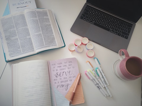 A neatly arranged workspace featuring an open black laptop, an open book with text, a notebook displaying a motivational quote written in neat handwriting, several colored marker pens, a stack of colorful washi tapes, and a pink and cream striped mug filled with a beverage.