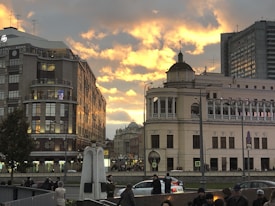A city street scene during sunset, featuring several people walking and taking photos. The skyline is dominated by older architectural buildings with ornate details, alongside modern blocks. The sky is dramatic with clouds lit by the orange and yellow hues of the setting sun.