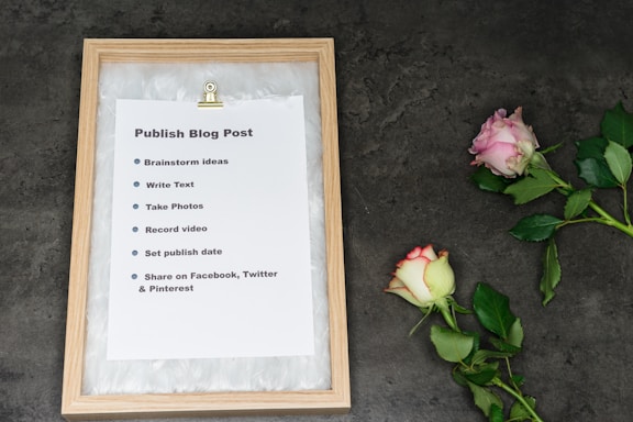 A framed checklist titled 'Publish Blog Post' is displayed on a textured, dark surface. The checklist includes steps such as brainstorming ideas, writing text, taking photos, recording video, setting a publish date, and sharing on social media platforms like Facebook, Twitter, and Pinterest. Next to the frame are two roses, one pink and one white with pink edges, accompanied by green leaves.