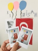 A hand holds two Polaroid photographs in front of a wall decorated with a large red flag featuring a white star and crescent, possibly the Turkish flag. Above the flag are cut-out paper balloons in yellow, pink, and blue, and a black-and-white portrait. The words '23 Nisan' are visible on the wall, formed with decorative letters, indicating a celebration, likely related to Turkish National Sovereignty and Children's Day.