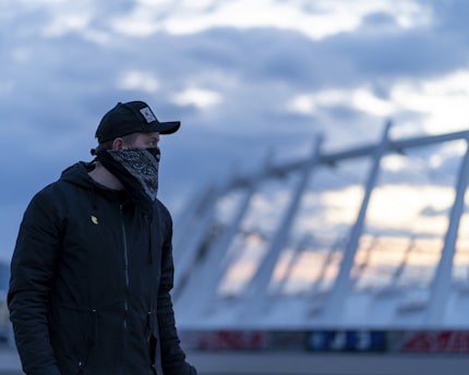 A person is wearing a black cap and a black jacket with a bandana covering their face. The background features a modern architectural structure with a cloudy sky during what appears to be twilight.