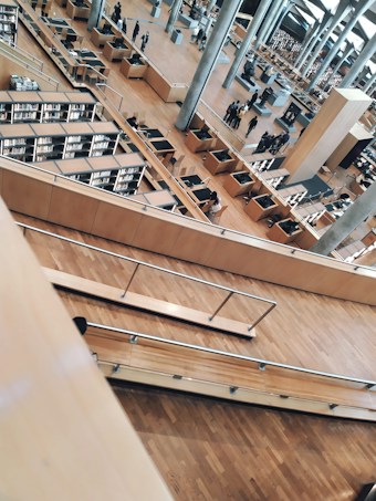 A spacious, modern library with multiple levels and rows of bookshelves. The open design features wood flooring, metallic railings, and several desks arranged for study. People can be seen walking around and engaging in activities in the background.