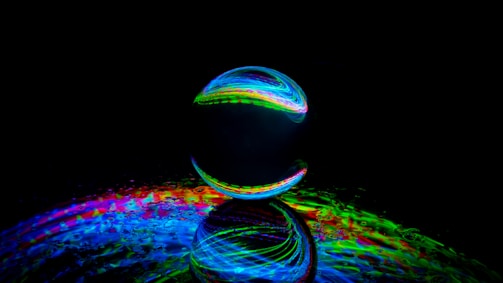 A reflective glass sphere is placed on a surface with a vibrant, multicolored light display. The light creates a rainbow effect, casting colorful patterns on the surface. The scene is set against a dark background, enhancing the luminous effect and creating an abstract, futuristic feel.