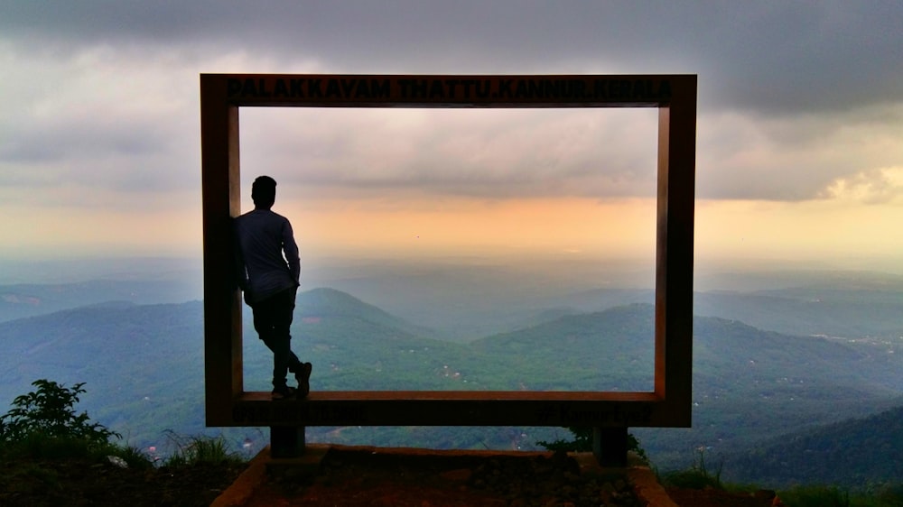 man in black jacket standing on brown wooden fence looking at mountains during daytime