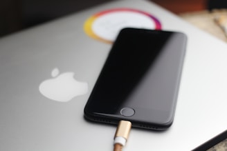 A sleek black smartphone is partially visible, resting on top of a silver laptop with a prominent white apple logo. The phone is connected to a charging cable with a gold-colored end. The scene is softly lit, with a blurred background, giving the image a minimalist and modern look.