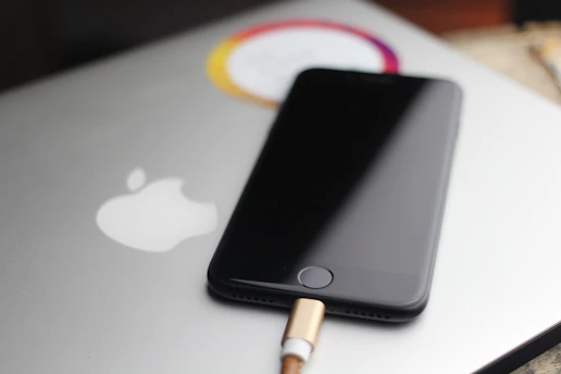 A sleek black smartphone is partially visible, resting on top of a silver laptop with a prominent white apple logo. The phone is connected to a charging cable with a gold-colored end. The scene is softly lit, with a blurred background, giving the image a minimalist and modern look.