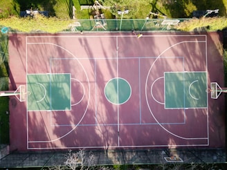 An aerial view of an outdoor basketball court featuring two large green squares on either end, separated by a red and brown surface with clearly marked white lines. The court is surrounded by grass and a fence on one side. Shadow patterns of trees are cast across the court, indicating a sunny day. There are no players on the court.