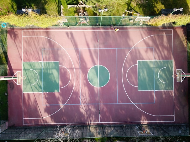 An aerial view of an outdoor basketball court featuring two large green squares on either end, separated by a red and brown surface with clearly marked white lines. The court is surrounded by grass and a fence on one side. Shadow patterns of trees are cast across the court, indicating a sunny day. There are no players on the court.