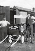 Two men stand beside a vintage milk cart labeled 'Royal Farm Dairy' with ornate design work. One man is dressed in a shirt with suspenders, while the other wears a vest and cap. The setting appears to be a farm with wooden structures and a sign reading 'Cows & Farms'. The image is in black and white, suggesting it is from an earlier era.