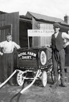Two men stand beside a vintage milk cart labeled 'Royal Farm Dairy' with ornate design work. One man is dressed in a shirt with suspenders, while the other wears a vest and cap. The setting appears to be a farm with wooden structures and a sign reading 'Cows & Farms'. The image is in black and white, suggesting it is from an earlier era.