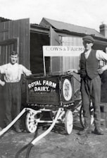 Two men stand beside a vintage milk cart labeled 'Royal Farm Dairy' with ornate design work. One man is dressed in a shirt with suspenders, while the other wears a vest and cap. The setting appears to be a farm with wooden structures and a sign reading 'Cows & Farms'. The image is in black and white, suggesting it is from an earlier era.