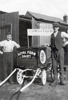 Two men stand beside a vintage milk cart labeled 'Royal Farm Dairy' with ornate design work. One man is dressed in a shirt with suspenders, while the other wears a vest and cap. The setting appears to be a farm with wooden structures and a sign reading 'Cows & Farms'. The image is in black and white, suggesting it is from an earlier era.