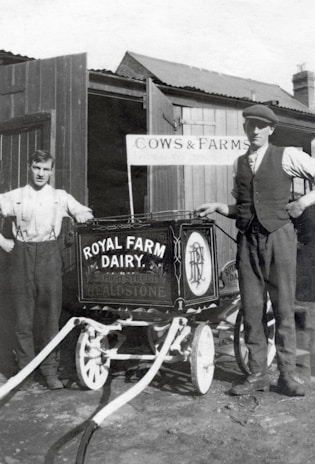 Two men stand beside a vintage milk cart labeled 'Royal Farm Dairy' with ornate design work. One man is dressed in a shirt with suspenders, while the other wears a vest and cap. The setting appears to be a farm with wooden structures and a sign reading 'Cows & Farms'. The image is in black and white, suggesting it is from an earlier era.
