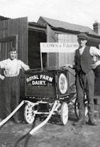 Two men stand beside a vintage milk cart labeled 'Royal Farm Dairy' with ornate design work. One man is dressed in a shirt with suspenders, while the other wears a vest and cap. The setting appears to be a farm with wooden structures and a sign reading 'Cows & Farms'. The image is in black and white, suggesting it is from an earlier era.