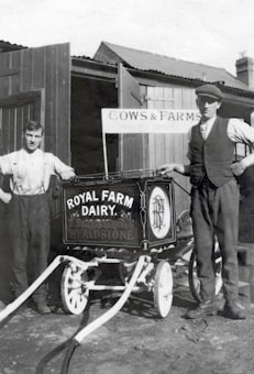 Two men stand beside a vintage milk cart labeled 'Royal Farm Dairy' with ornate design work. One man is dressed in a shirt with suspenders, while the other wears a vest and cap. The setting appears to be a farm with wooden structures and a sign reading 'Cows & Farms'. The image is in black and white, suggesting it is from an earlier era.