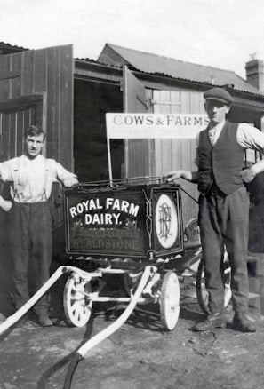 Two men stand beside a vintage milk cart labeled 'Royal Farm Dairy' with ornate design work. One man is dressed in a shirt with suspenders, while the other wears a vest and cap. The setting appears to be a farm with wooden structures and a sign reading 'Cows & Farms'. The image is in black and white, suggesting it is from an earlier era.