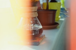 A glass coffee maker with wooden accents filled with coffee sits on a coaster. The background includes blurred potted plants. Light leaks across the image create a warm, nostalgic effect.