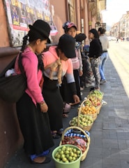 Several women dressed in traditional clothing are standing on a street, examining baskets of fruits arranged on the pavement. The fruits include apples, pears, strawberries, and other produce. The scene appears to be a market setting with posters on the wall behind them and people walking on the street.