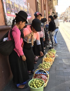 Several women dressed in traditional clothing are standing on a street, examining baskets of fruits arranged on the pavement. The fruits include apples, pears, strawberries, and other produce. The scene appears to be a market setting with posters on the wall behind them and people walking on the street.