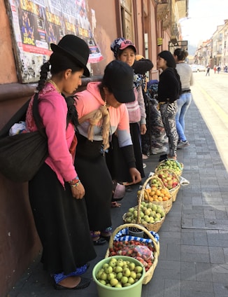 Several women dressed in traditional clothing are standing on a street, examining baskets of fruits arranged on the pavement. The fruits include apples, pears, strawberries, and other produce. The scene appears to be a market setting with posters on the wall behind them and people walking on the street.