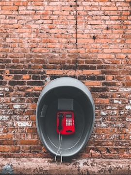 A red public telephone is mounted inside a protective metal casing against an old brick wall. The wall has a rustic appearance, with a combination of orange and brown brick colors. The phone is suspended by a metal cord and contrasts sharply with the surrounding industrial textures.