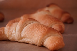 Several freshly baked croissants with a golden-brown crust are resting on a sheet of parchment paper. The focus is on the front croissant, showcasing its flaky, buttery layers and textured surface.