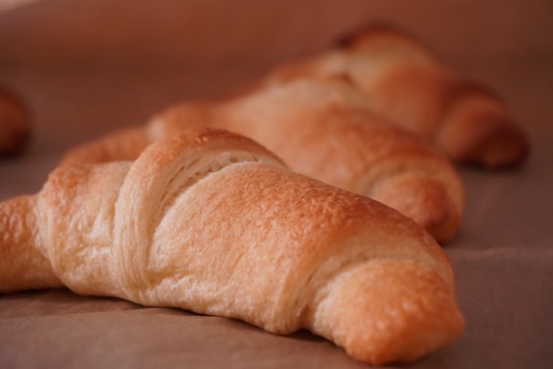Several freshly baked croissants with a golden-brown crust are resting on a sheet of parchment paper. The focus is on the front croissant, showcasing its flaky, buttery layers and textured surface.