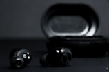 A pair of black wireless earbuds is positioned in front of a black charging case on a dark background. The earbuds show metallic accents and small gold connectors for charging.
