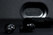 A pair of black wireless earbuds is positioned in front of a black charging case on a dark background. The earbuds show metallic accents and small gold connectors for charging.