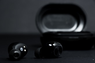 A pair of black wireless earbuds is positioned in front of a black charging case on a dark background. The earbuds show metallic accents and small gold connectors for charging.