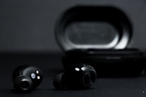 A pair of black wireless earbuds is positioned in front of a black charging case on a dark background. The earbuds show metallic accents and small gold connectors for charging.