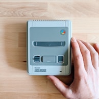 A hand is gently placed on a gray Super Nintendo Entertainment System console, positioned on a wooden surface. The console features a power button, eject button, and reset button, along with a colorful logo.