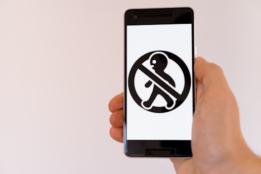 A hand is holding a smartphone displaying a banned pedestrian symbol in black on a white background. The symbol consists of a person walking, crossed out within a circle, typically used to indicate a prohibition.