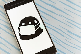 A smartphone displays a black and white illustration of a masked face with sweat droplets. The phone is placed on a light blue surface with diagonal, irregular lines resembling raindrops or waves.