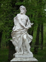 A classical white marble statue depicting a muscular male figure, standing on a pedestal in a natural setting. The figure is draped in flowing cloth, with an expression of determination. The background is lush with green trees, indicating a peaceful outdoor environment.