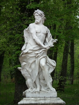 A classical white marble statue depicting a muscular male figure, standing on a pedestal in a natural setting. The figure is draped in flowing cloth, with an expression of determination. The background is lush with green trees, indicating a peaceful outdoor environment.