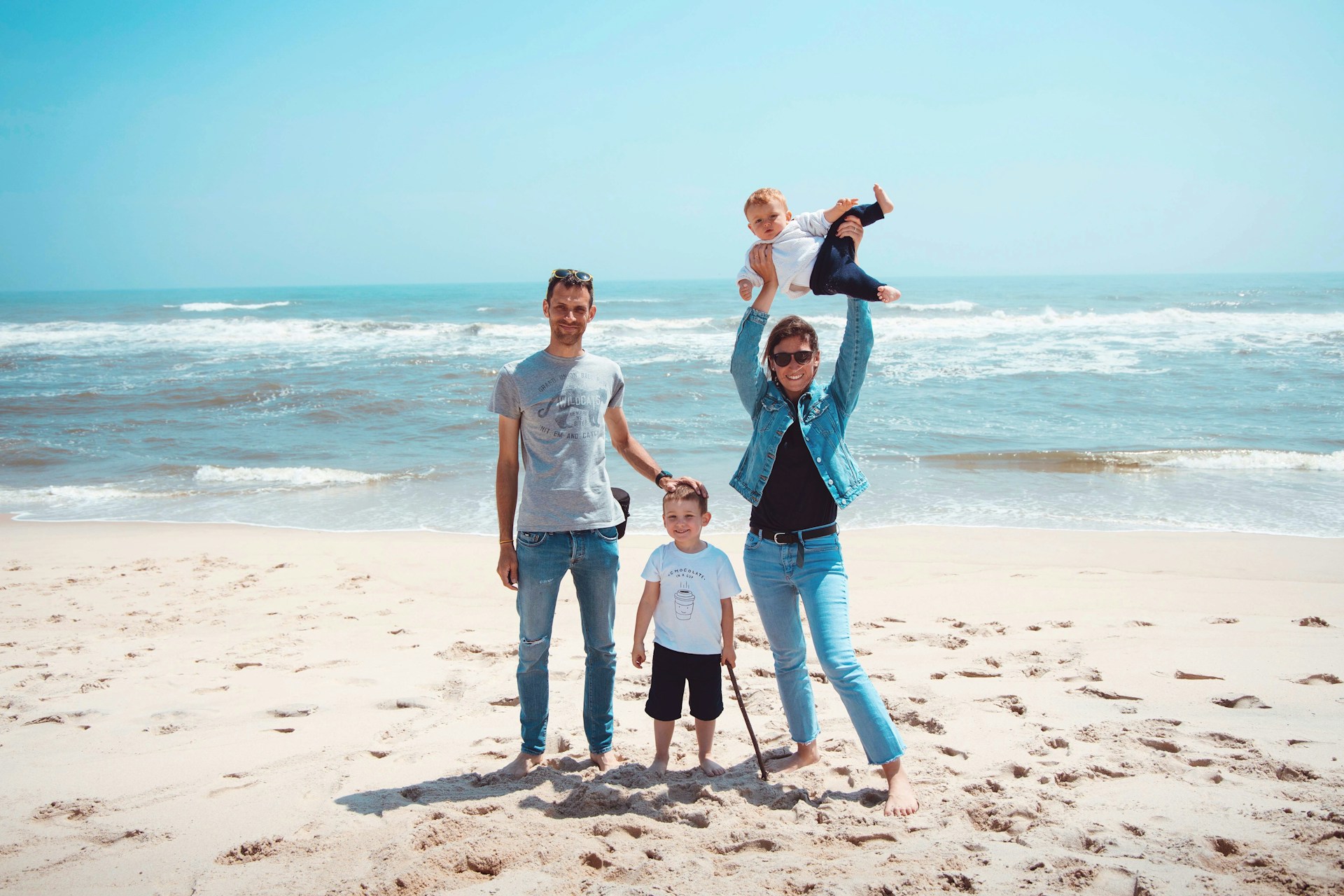 A family stands on a sandy beach with the ocean waves in the background. The sky is clear and blue, suggesting a sunny day. The group includes two adults and two children, smiling and posing happily.
