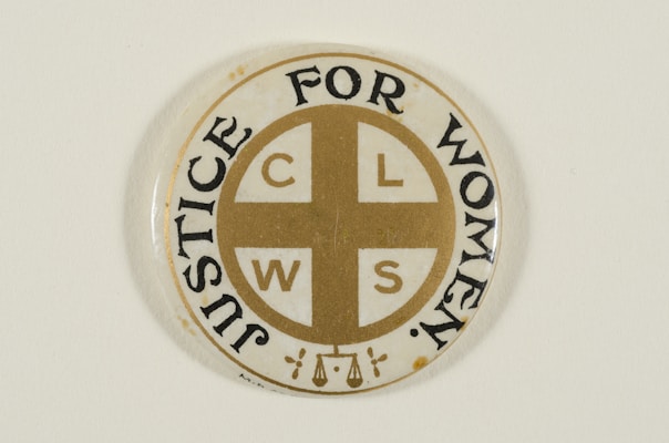 A circular pin badge with a gold cross dividing the letters 'C', 'L', 'W', 'S' in each quadrant. Surrounding the cross, the words 'Justice For Women' are printed in bold black text. The badge background is predominantly white, with a subtle decorative element at the bottom.