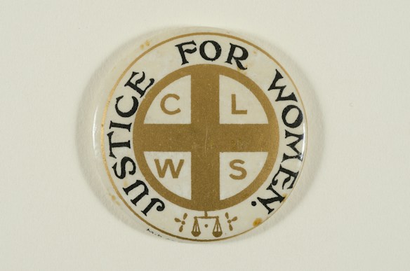 A circular pin badge with a gold cross dividing the letters 'C', 'L', 'W', 'S' in each quadrant. Surrounding the cross, the words 'Justice For Women' are printed in bold black text. The badge background is predominantly white, with a subtle decorative element at the bottom.