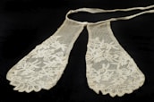 A delicate, vintage lace accessory featuring intricate floral patterns and a soft texture. The lace appears to lie on a flat, dark surface and is cream in color.