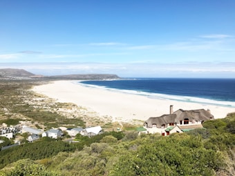 A scenic coastal landscape featuring a vast, white sandy beach that stretches along the deep blue ocean. In the foreground, there are green, bushy areas and a few houses, including a prominent thatched-roof cottage. The sky is clear with a few clouds, and mountains can be seen in the background.